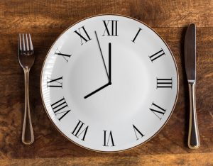 Eight hour feeding window concept or breakfast time with clock on plate and knife and fork on wooden table, overhead view-img-blog