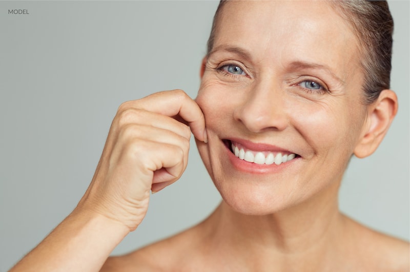 Beautiful aging woman gently pulling at her cheek skin with a smile on her face.